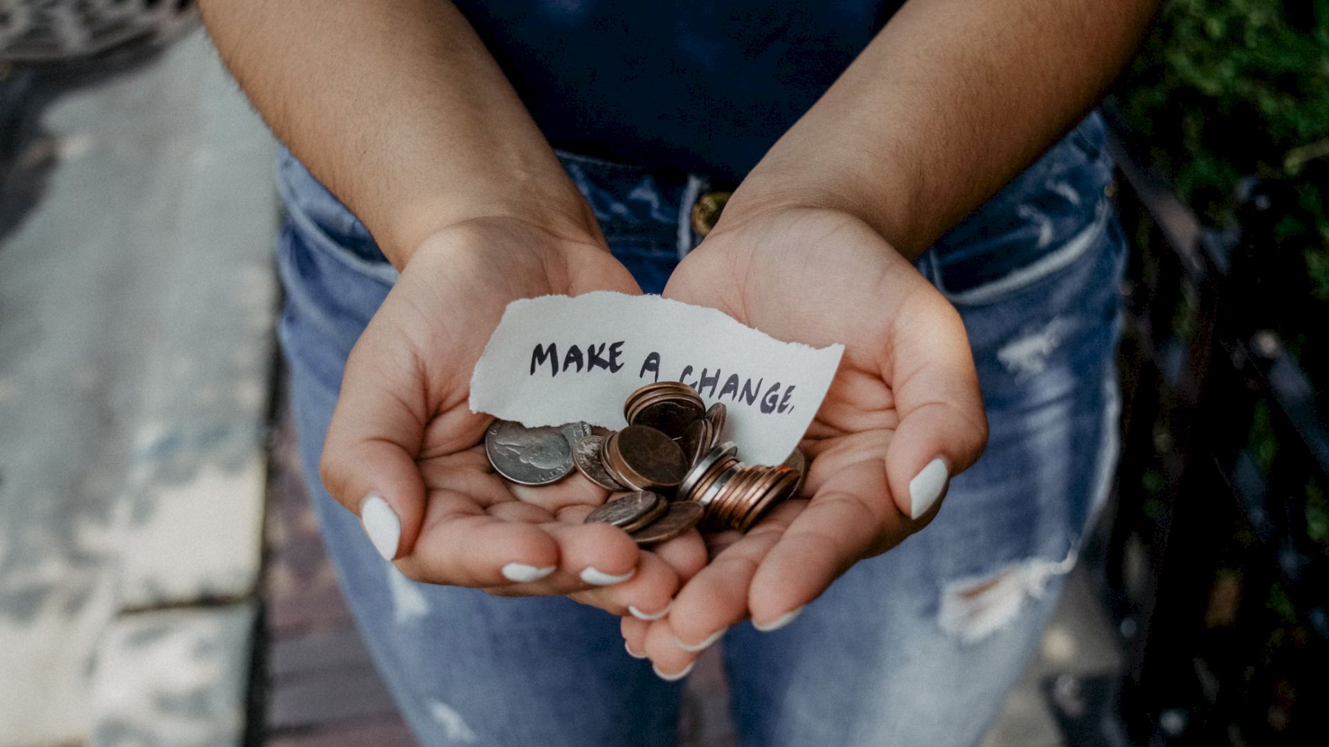 Two hands holding a small pile of coins and a note that reads "MAKE A CHANGE" on a piece of paper.