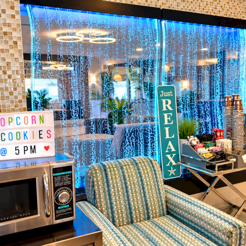 A cozy lounge photo booth with blue cascading lights, popcorn & cookies sign, a &ldquo;Just Relax&rdquo; column, and snack cart and comfy chairs.