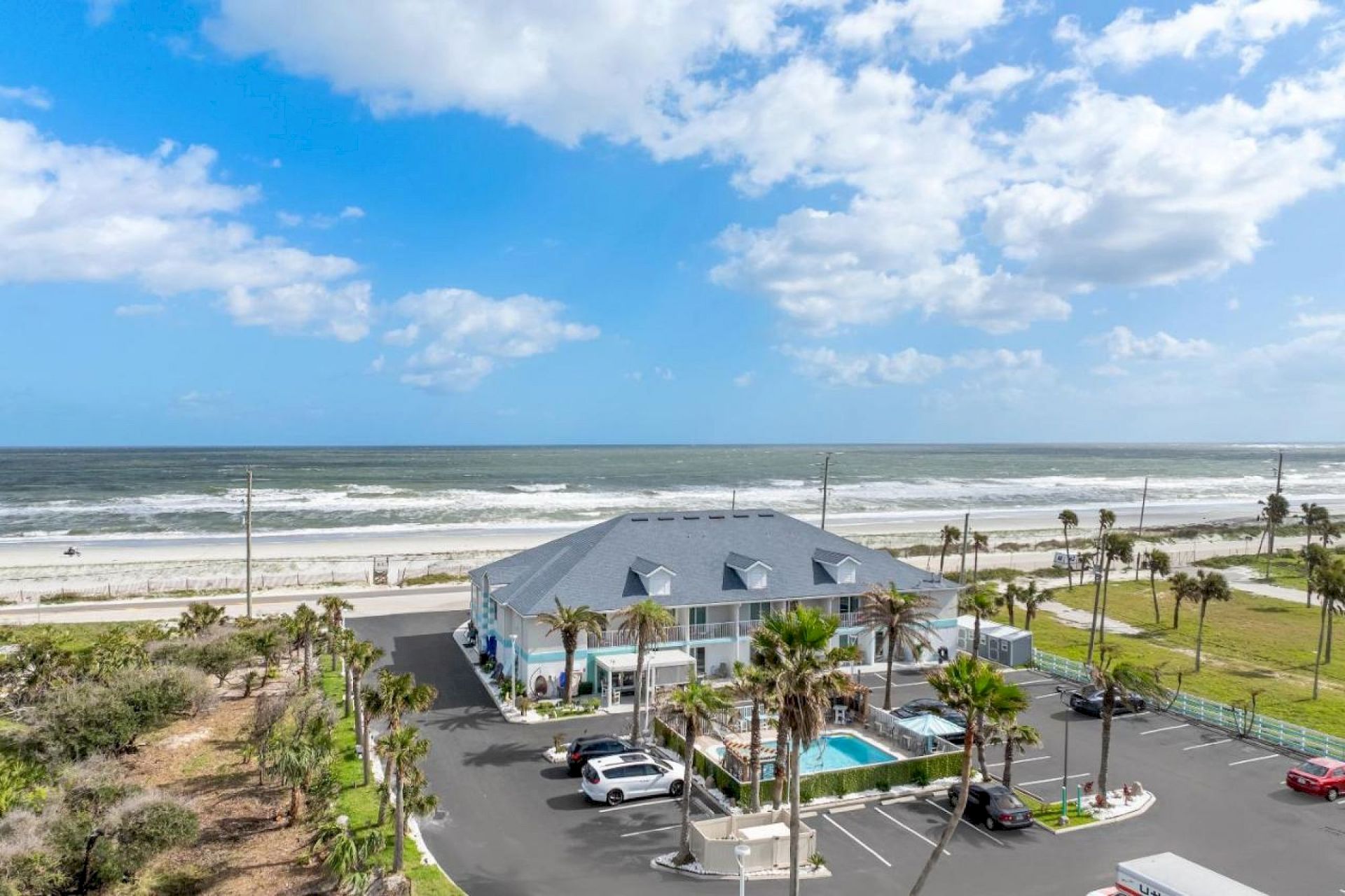 A coastal scene shows a building with parked cars, a pool, and a beach with waves under a partly cloudy sky in the background.