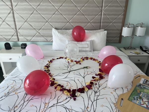 A bed decorated with a heart made of rose petals and surrounded by red, pink, and white balloons, with a single rose in the center.