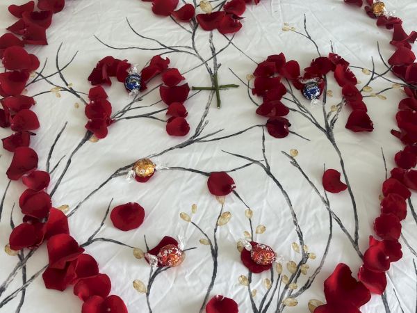 Red rose petals form a heart, cross, and "M" shapes on a white bed, adorned with wrapped chocolate candies scattered inside.