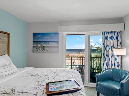 A bright hotel bedroom with a bed, blue chair and lamp, and a balcony window showing an ocean view.