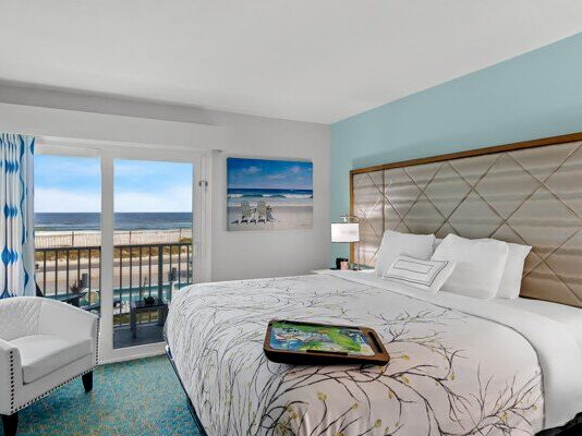A bright hotel bedroom with a large bed and ocean-view balcony, plus framed seascape art on the wall.