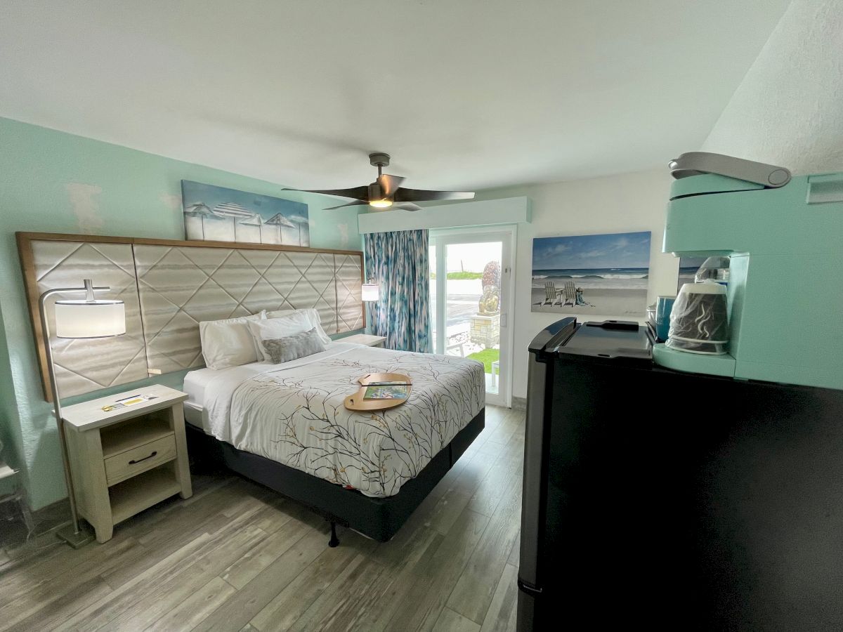 A bright seaside-themed bedroom with a bed, ceiling fan, ocean artwork, curtains, and a countertop coffee machine near the window.