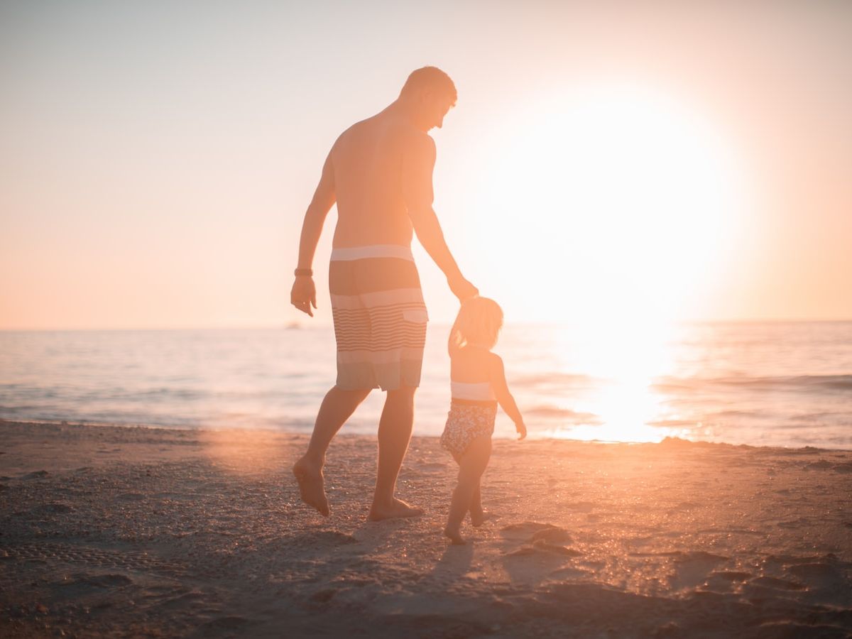 A man and child walk hand in hand on a beach at sunset, with the sun setting over the ocean in the background.