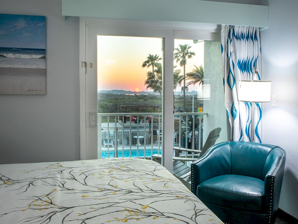 A cozy room features a bed, teal chair, and window with a sunset view. Outside, palm trees and a pool are visible through glass doors.
