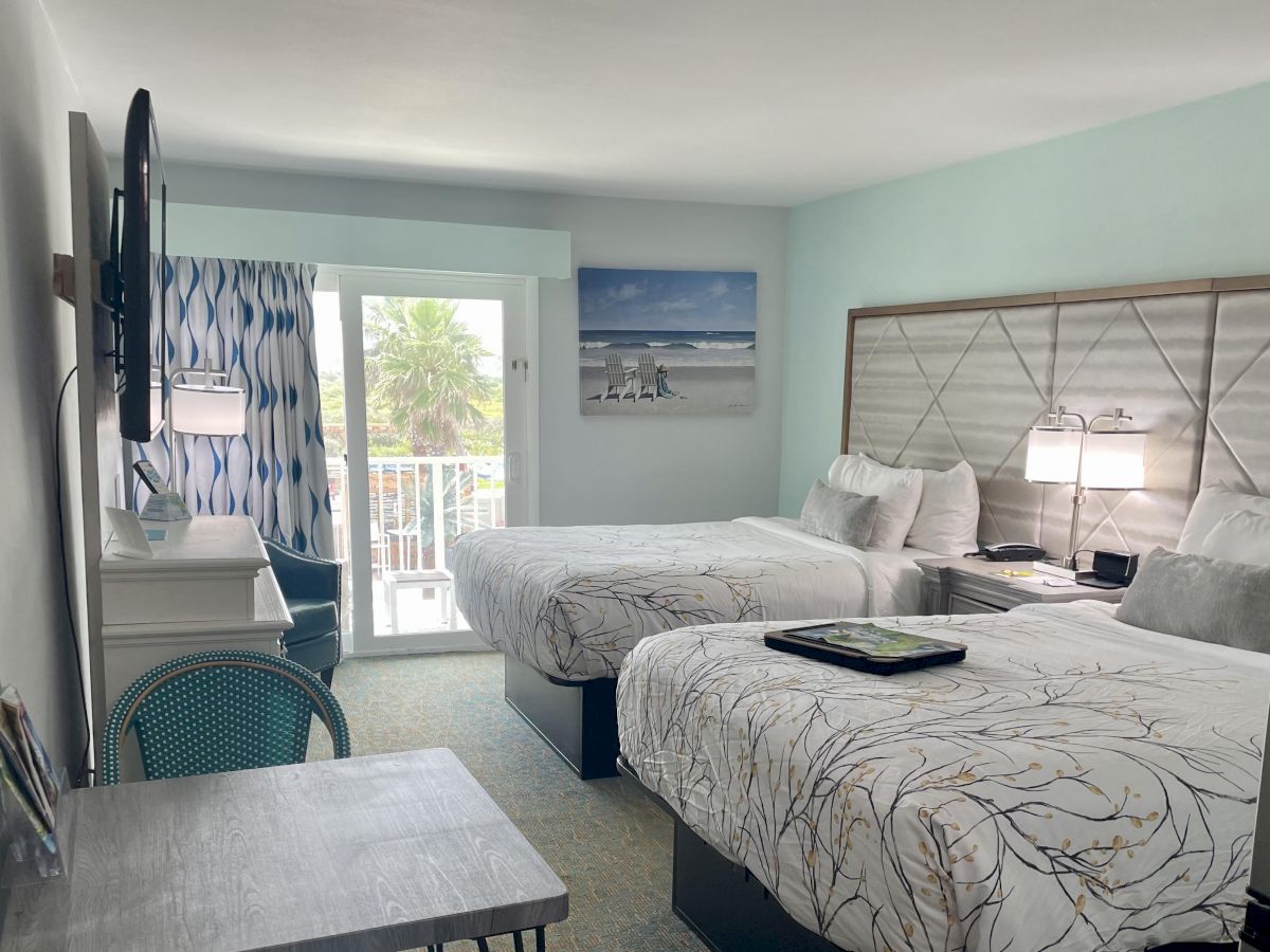 A hotel room features two beds, beach-themed decor, a TV, and a small desk, with a balcony view of palm trees and the ocean.